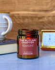 Lit candle labeled The Autumn Chapter with blurred background of book, mug and clock