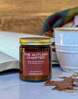 Candle labeled The Autumn Chapter with background of book, mug and plaid blanket