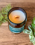 Overhead of lit candle labeled The Forager's Hearth on wooden background surrounded by herbs