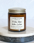A jar candle with a label that reads 'To The Stars Who Listen' on a white marble coaster. The jar is transparent amber with a gold lid.