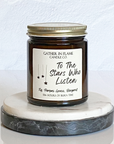 A jar candle with a label that reads 'To The Stars Who Listen' on a white marble coaster. The jar is transparent amber with a gold lid.
