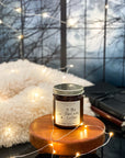 A jar candle with a label that reads 'To The Stars Who Listen' on a wooden surface with a dark window, leather books, fairy lights, and furry blanket in the background.