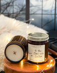 A jar candle with a label that reads 'To The Stars Who Listen' on a wooden surface next to a candle showing a smooth surface and a wood wick with a dark window, leather books, fairy lights, and furry blanket in the background.