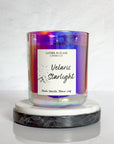 A multicolored, star-patterned candle in an iridescent glass jar, with a label reading 'Velaris Starlight'. The candle is placed on a white marble surface.