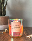 A multicolored, star-patterned candle in an iridescent glass jar, with a label reading 'Velaris Starlight'. The candle is placed on a wooden surface with matches and a wick trimmer and plant in the background.