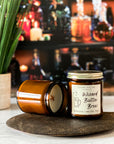 Jar candle with a cream label that reads 'Wizard Butter Brew' with a illustration of a mug next to candle showing creamy surface and wood x wick, on a wood surface with a plant and bottles in the background.