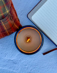 Topdown view of lit candle with wood wick with a notebook and plaid flannel shirt on a blue surface