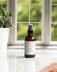 A brown amber spray bottle labeled 'Cozy Cashmere' by Gather In Flame Candle Co. on a white marble surface with a window and plant in the background.