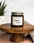 3.6oz candle jar of SSugared Citrus on wooden pedestal next to plant