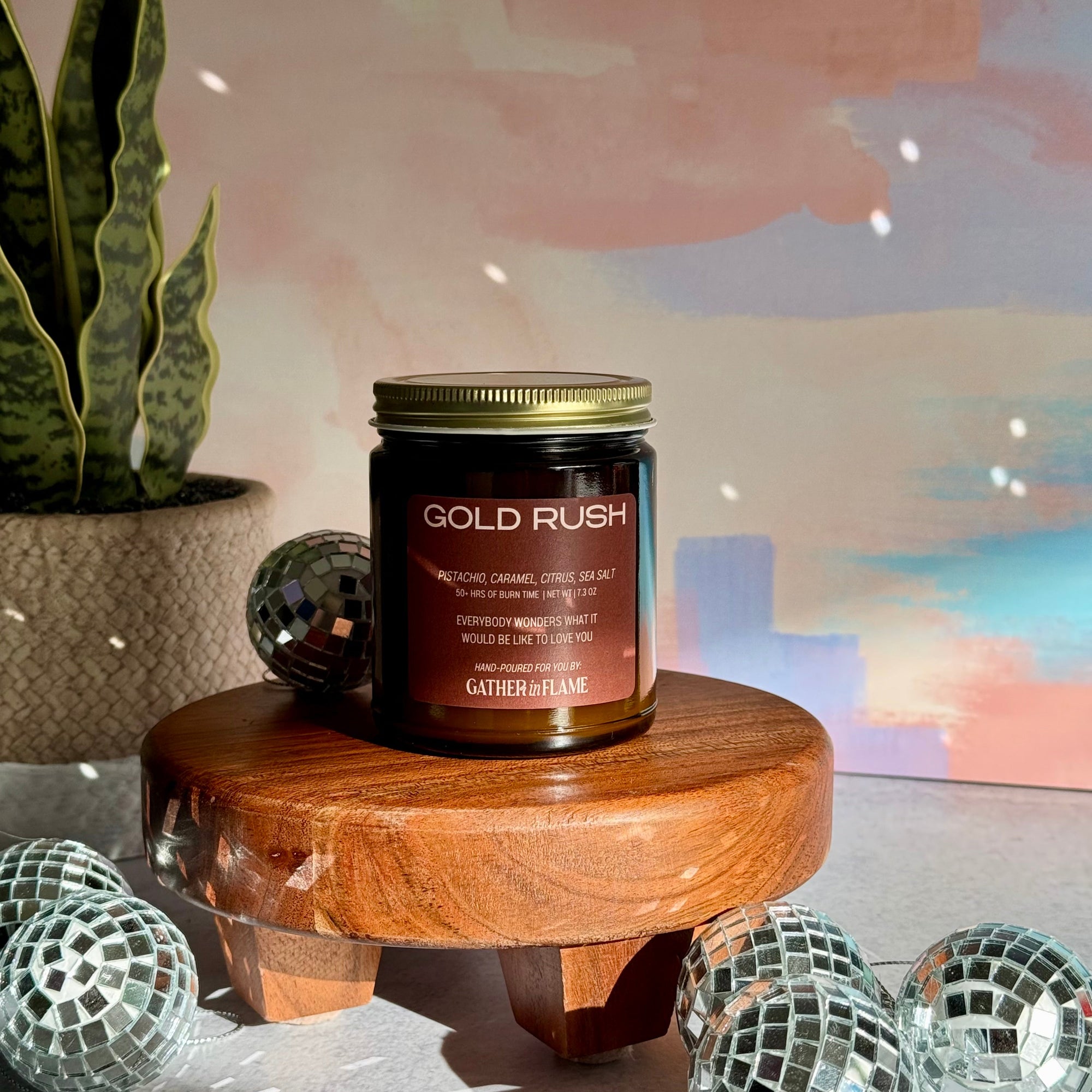 Candle labeled 'Gold Rush' on a wooden stand with decorative items and a plant in the background.