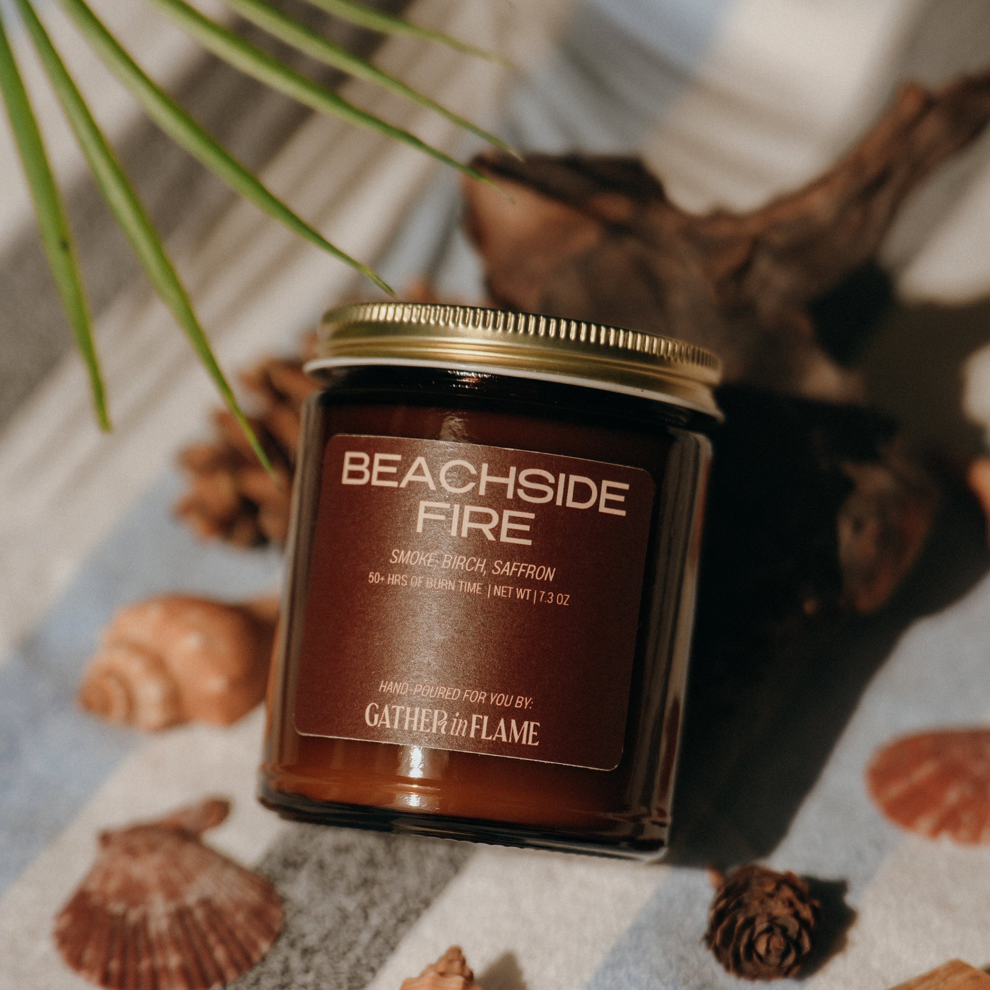 Candle labeled 'Beachside Fire' by Gather of Flame on a surface with natural elements like shells and wood.