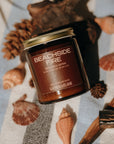 Candle jar labeled 'Beachside Fire' on a surface with seashells and driftwood.