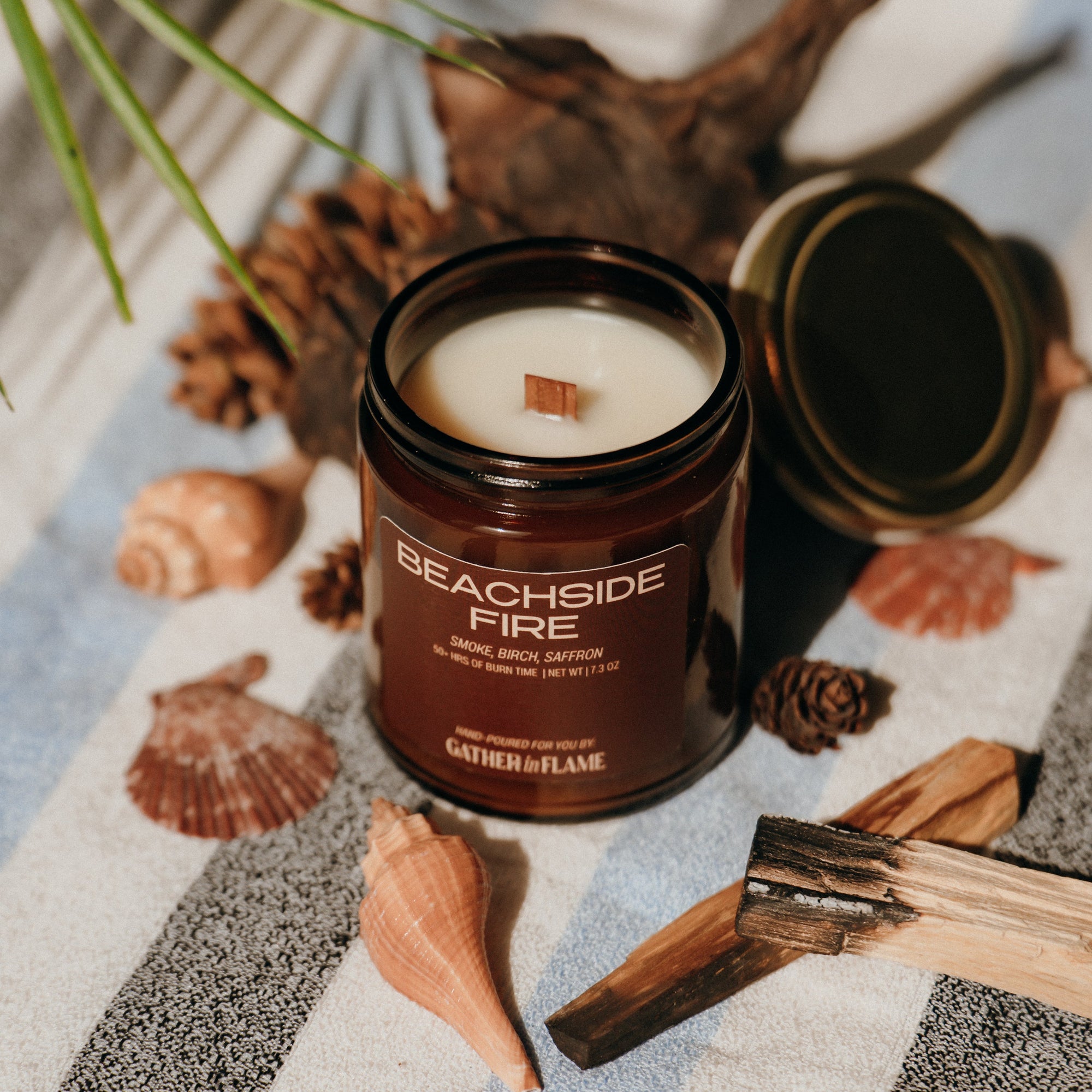 Candle labeled 'Beachside Fire' on a striped fabric with shells and wood, evoking a beachside theme.
