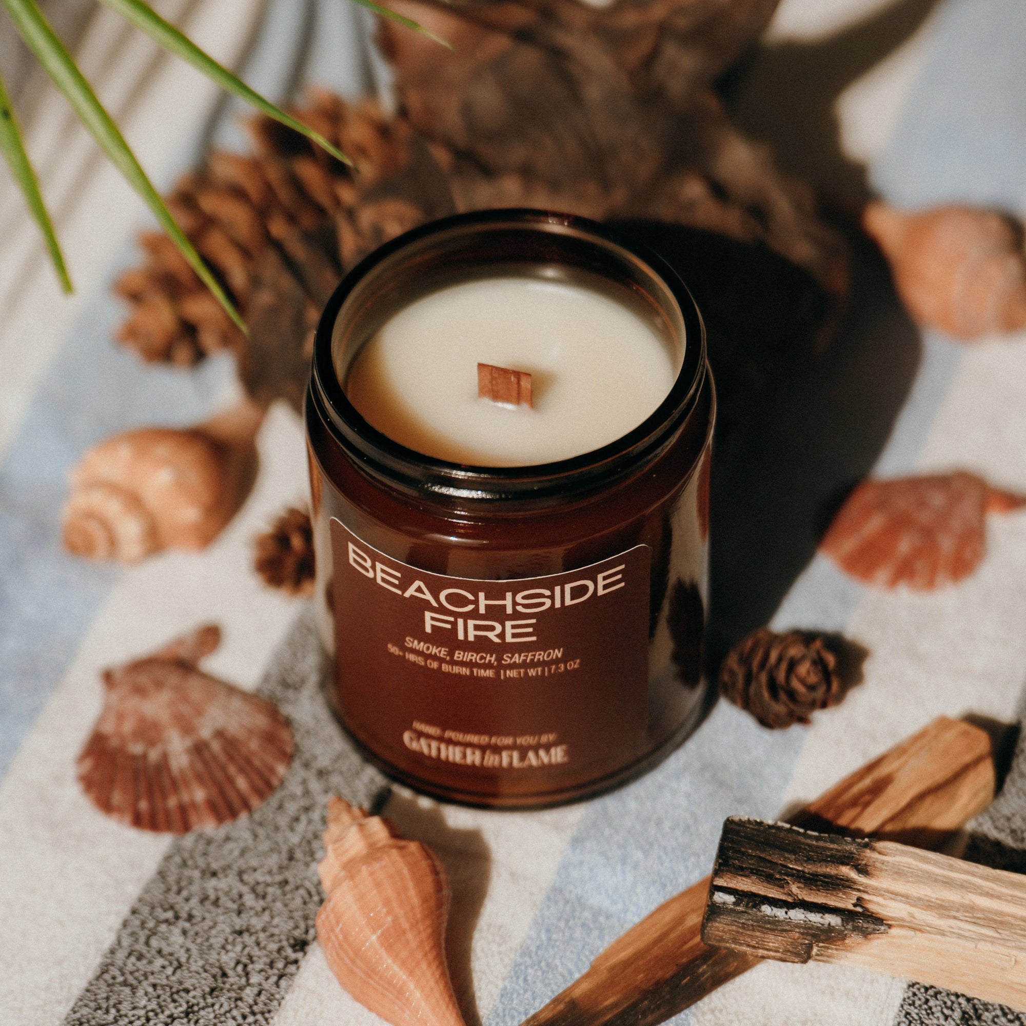 Candle labeled 'Beachside Fire' on a striped fabric with seashells and driftwood.