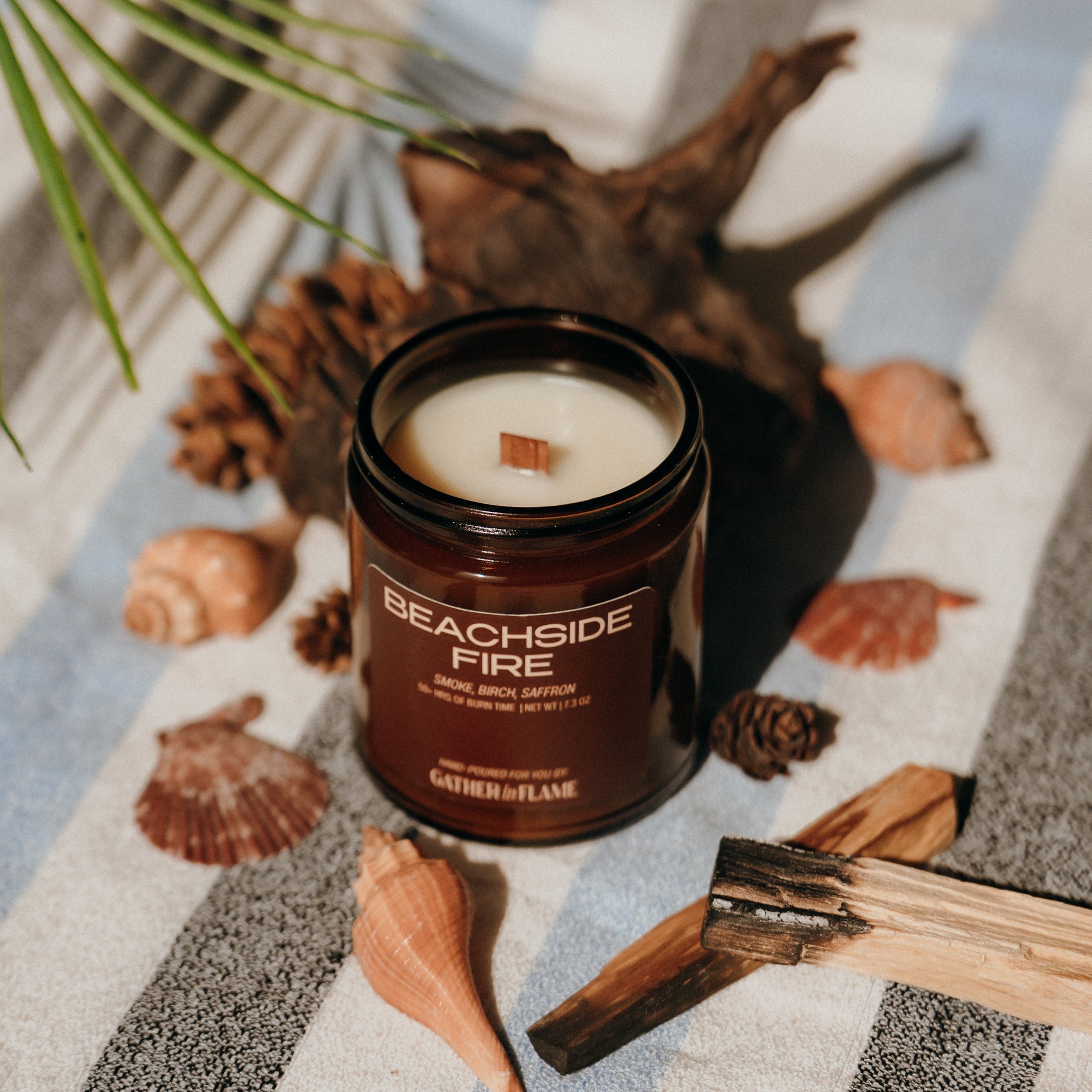 Candle labeled 'Beachside Fire' on a striped surface with shells and a burnt wood.