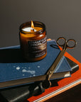 Candle in a brown glass jar on a book with a pair of scissors next to it against a dark background