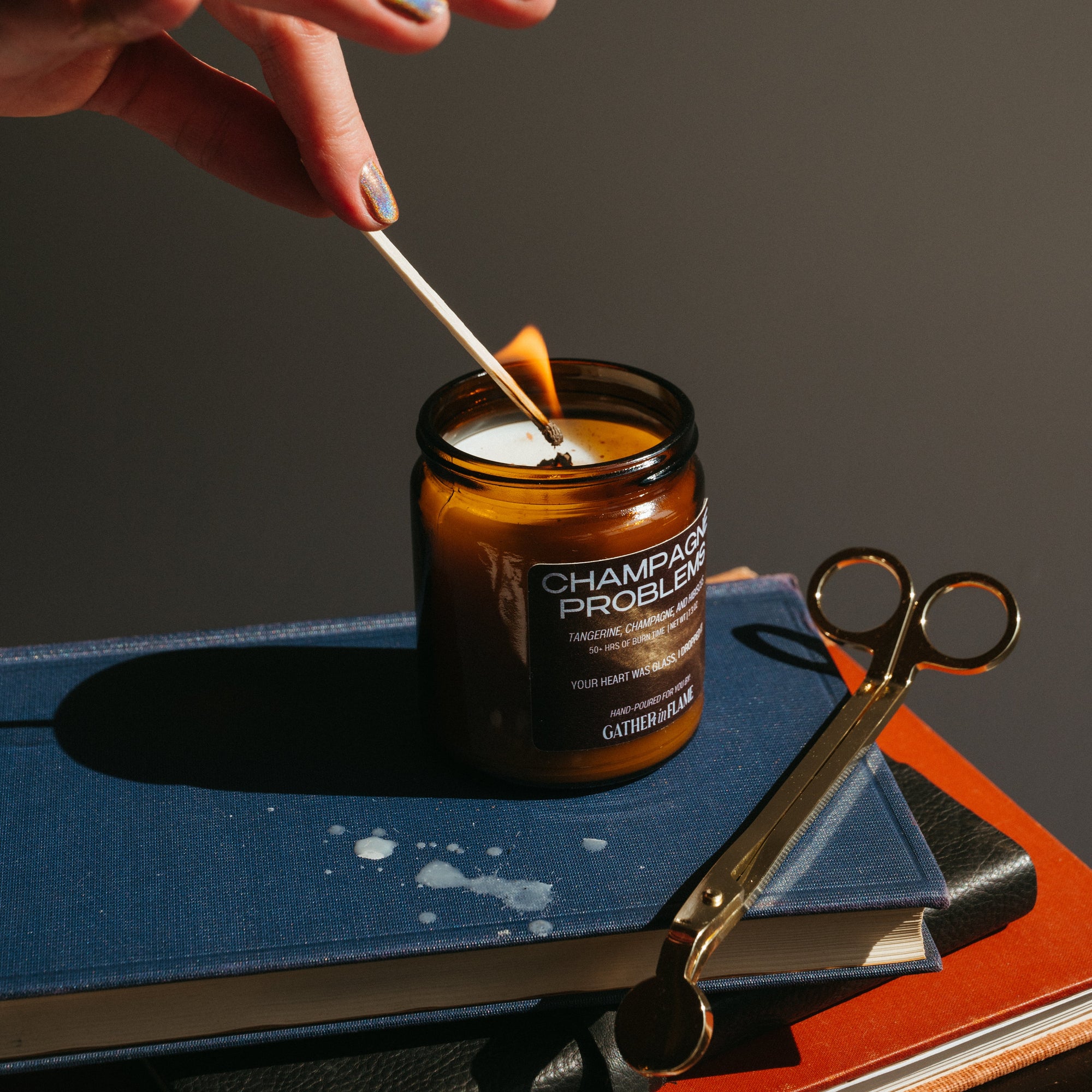 Person lighting a candle with a match, surrounded by scissors and a book on a dark surface.
