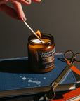Person lighting a candle with a match, surrounded by scissors and a book on a dark surface.
