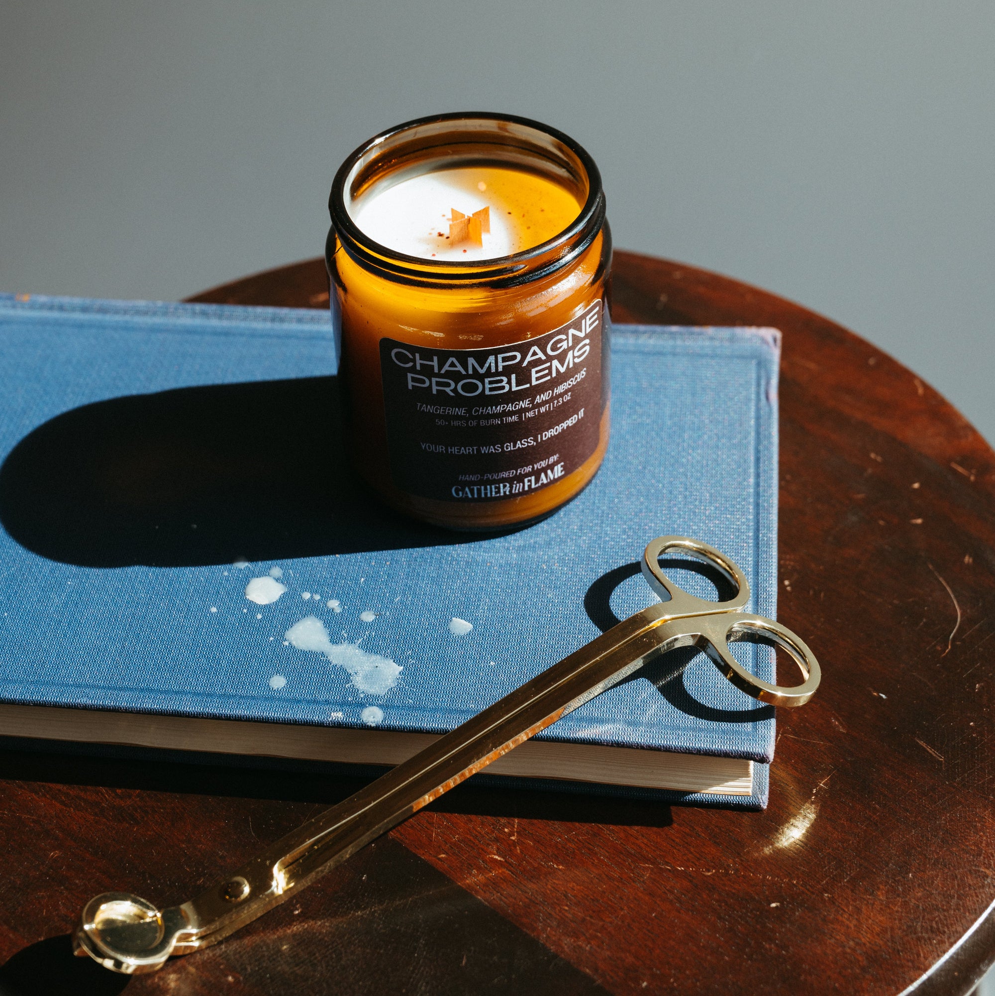 Candle labeled 'Champagne Problem' on a book with gold wick trimmers on a wooden surface