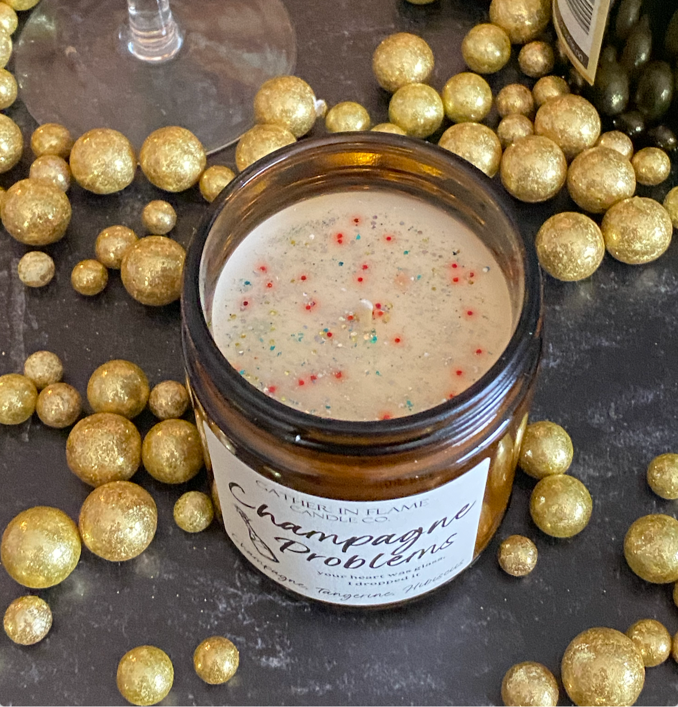 A jar candle with a label reading 'Champagne Problems' surrounded by gold metallic balls and showing eco-glitter on top