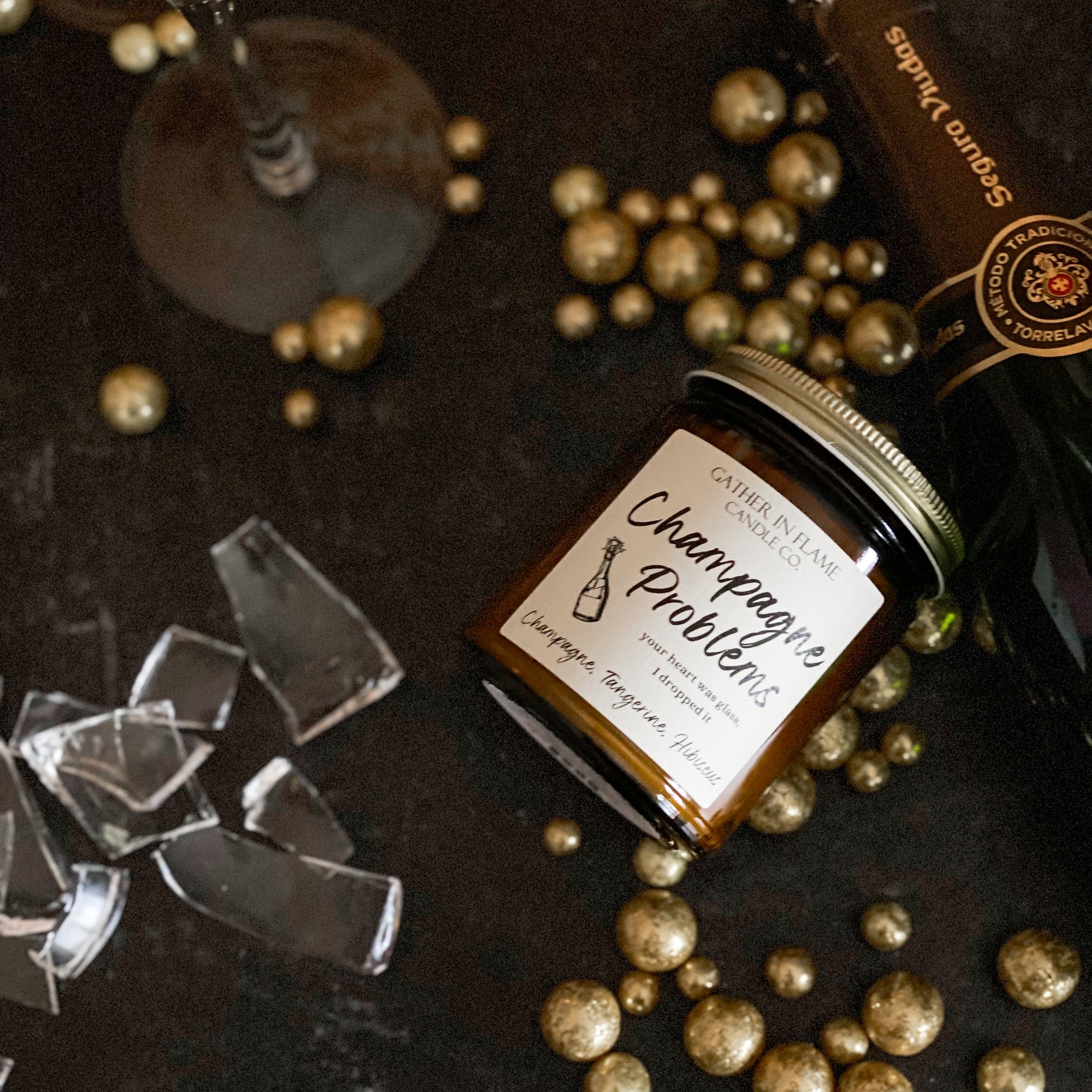 A jar candle with a label reading 'Champagne Problems' surrounded by broken glass and gold metallic balls