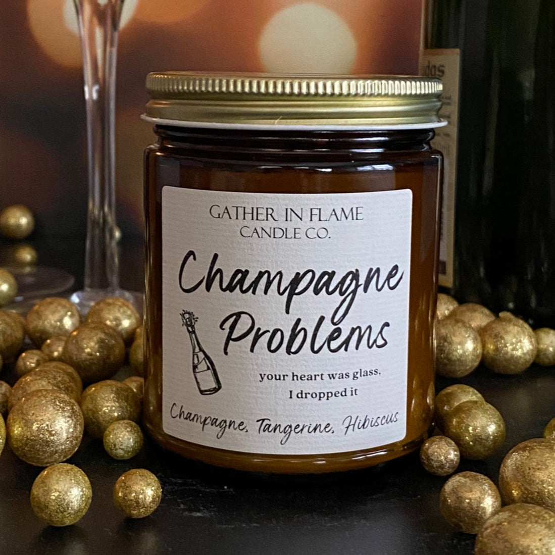 A jar candle with a label reading 'Champagne Problems' surrounded by gold metallic balls