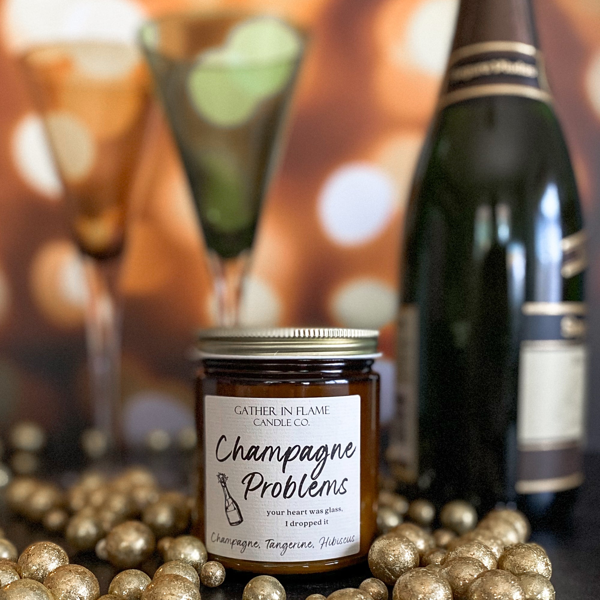 A jar candle with a label reading 'Champagne Problems' surrounded by gold metallic balls, a bottle of champagne, and champagne flutes.