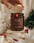 Hand holding a jar of 'City of Roses' candle with decorative elements on a soft background