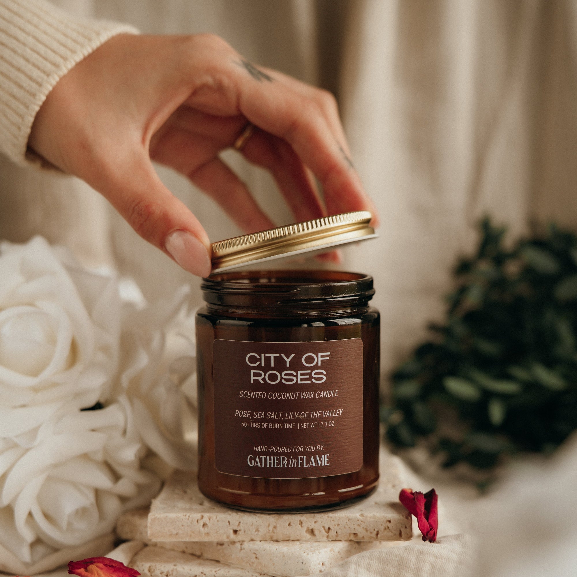 Hand opening a candle jar labeled 'City of Roses' with decorative elements in the background.