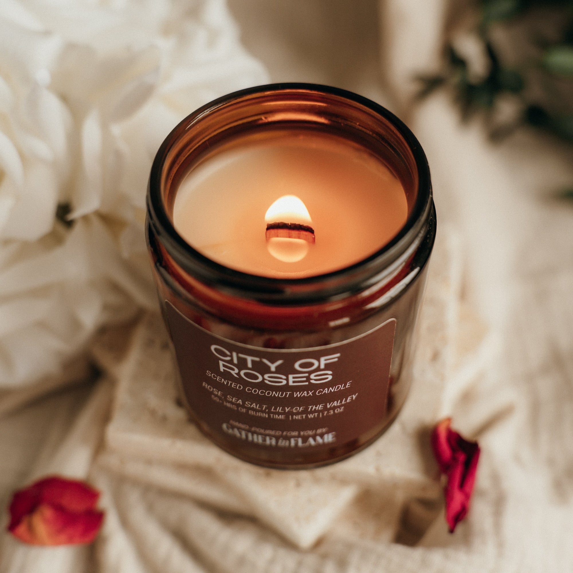 Candle in a brown jar labeled 'City of Roses' on a soft fabric background with red petals.