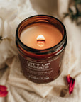 Candle in a brown jar labeled 'City of Roses' on a soft fabric background with red petals.
