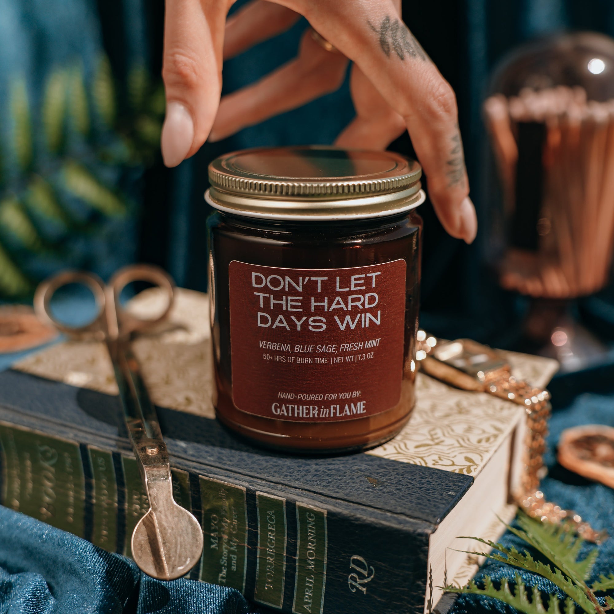 Hand touching the lid of a candle jar labeled 'Don't Let the Hard Days Win', surrounded by decorative items.