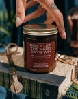 Hand touching the lid of a candle jar labeled 'Don't Let the Hard Days Win', surrounded by decorative items.