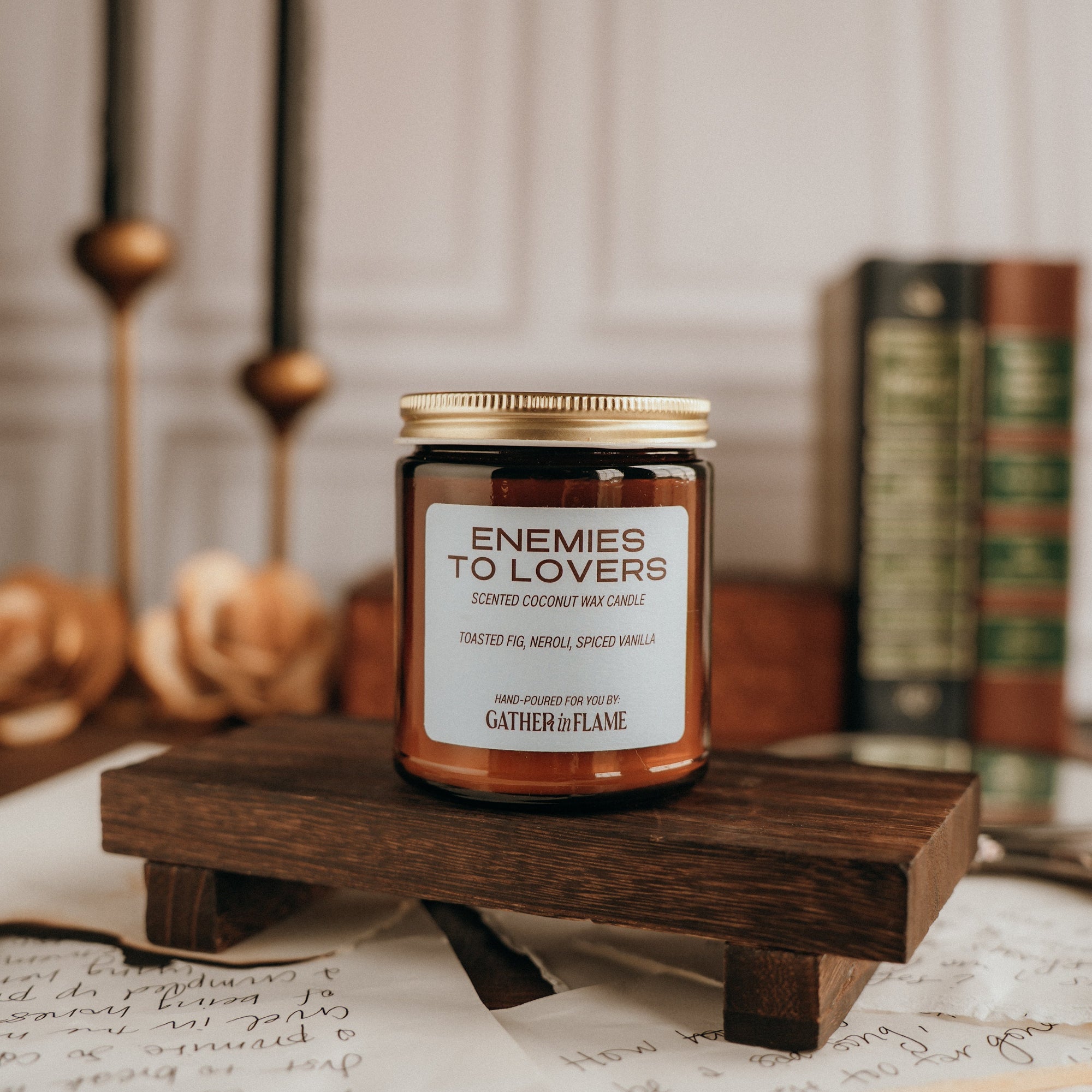 Candle labeled 'Enemies to Lovers' on a wooden stand with candles and books in the background.