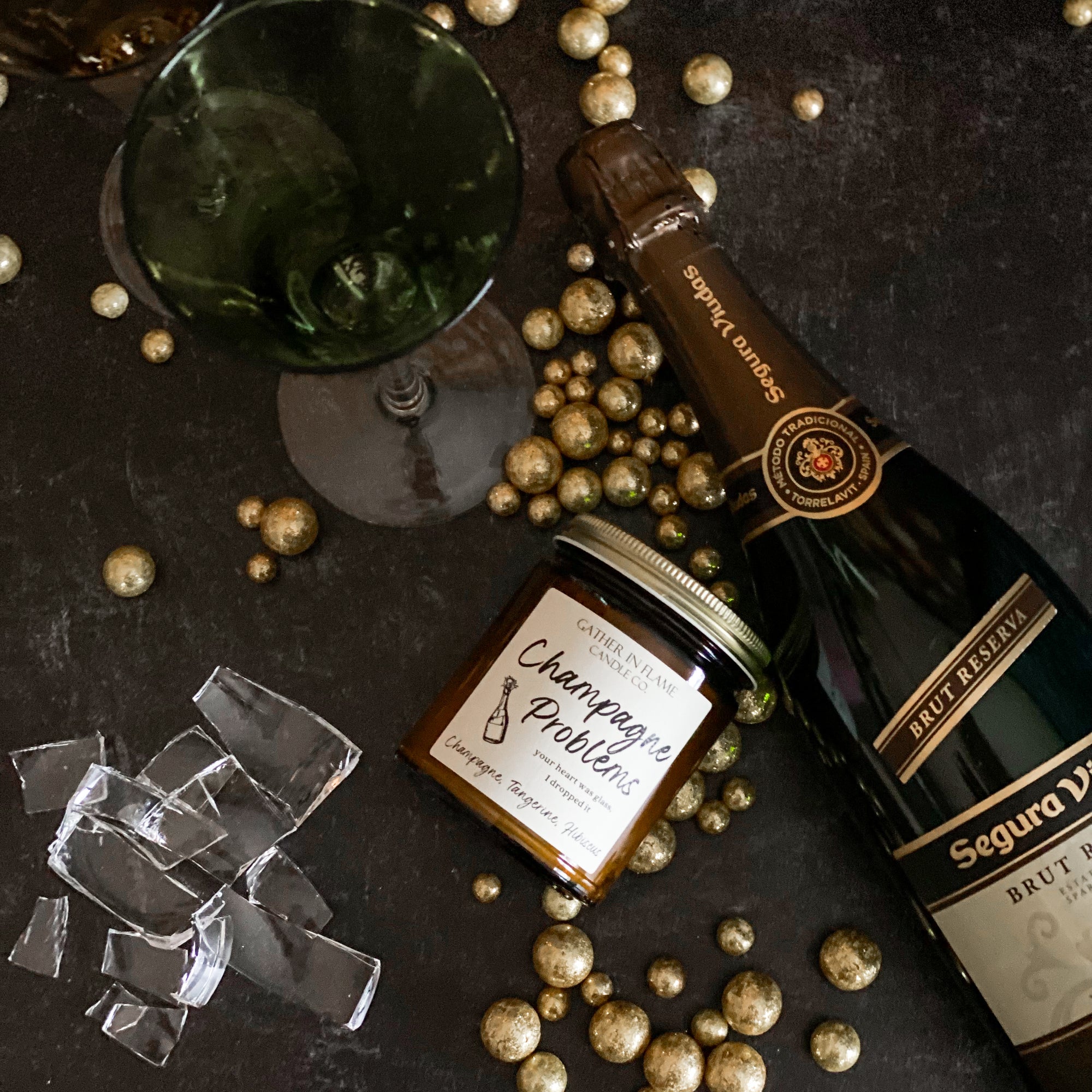A jar candle with a label reading &#39;Champagne Problems&#39; surrounded by gold metallic balls , broken glass, champagne bottle and champagne flutes