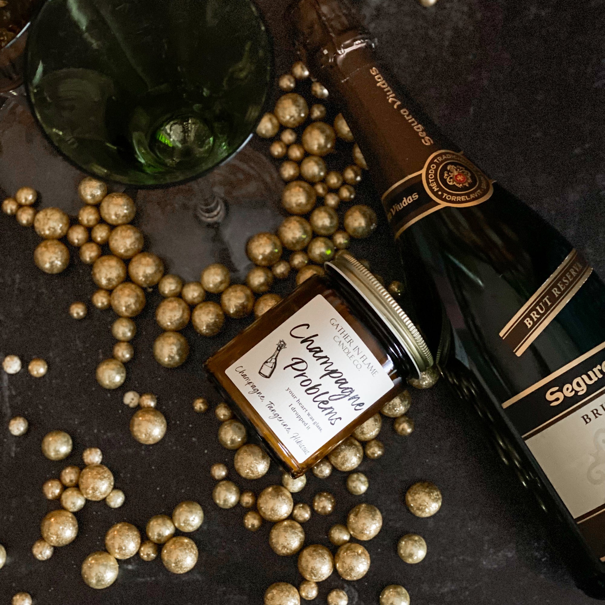 A jar candle with a label reading &#39;Champagne Problems&#39; surrounded by gold metallic balls, champagne bottle and champagne flutes