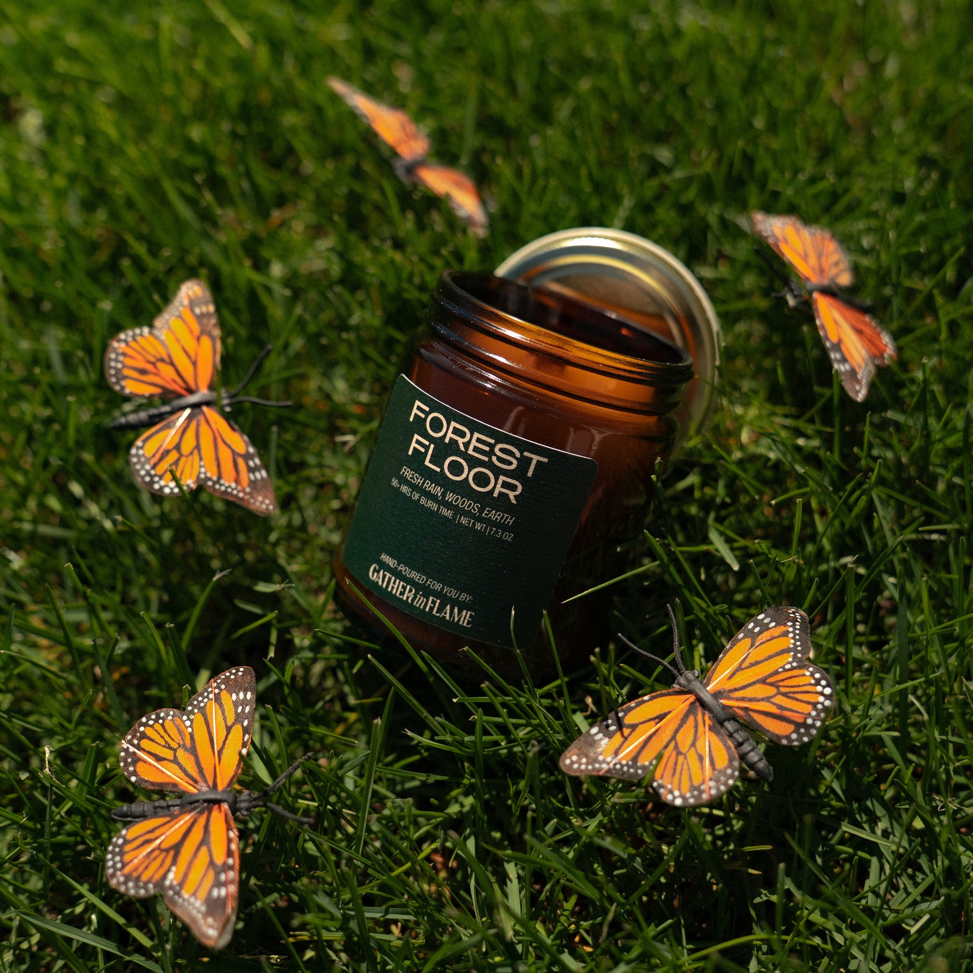 Jar of 'Forest Floor' candle surrounded by monarch butterflies on grass