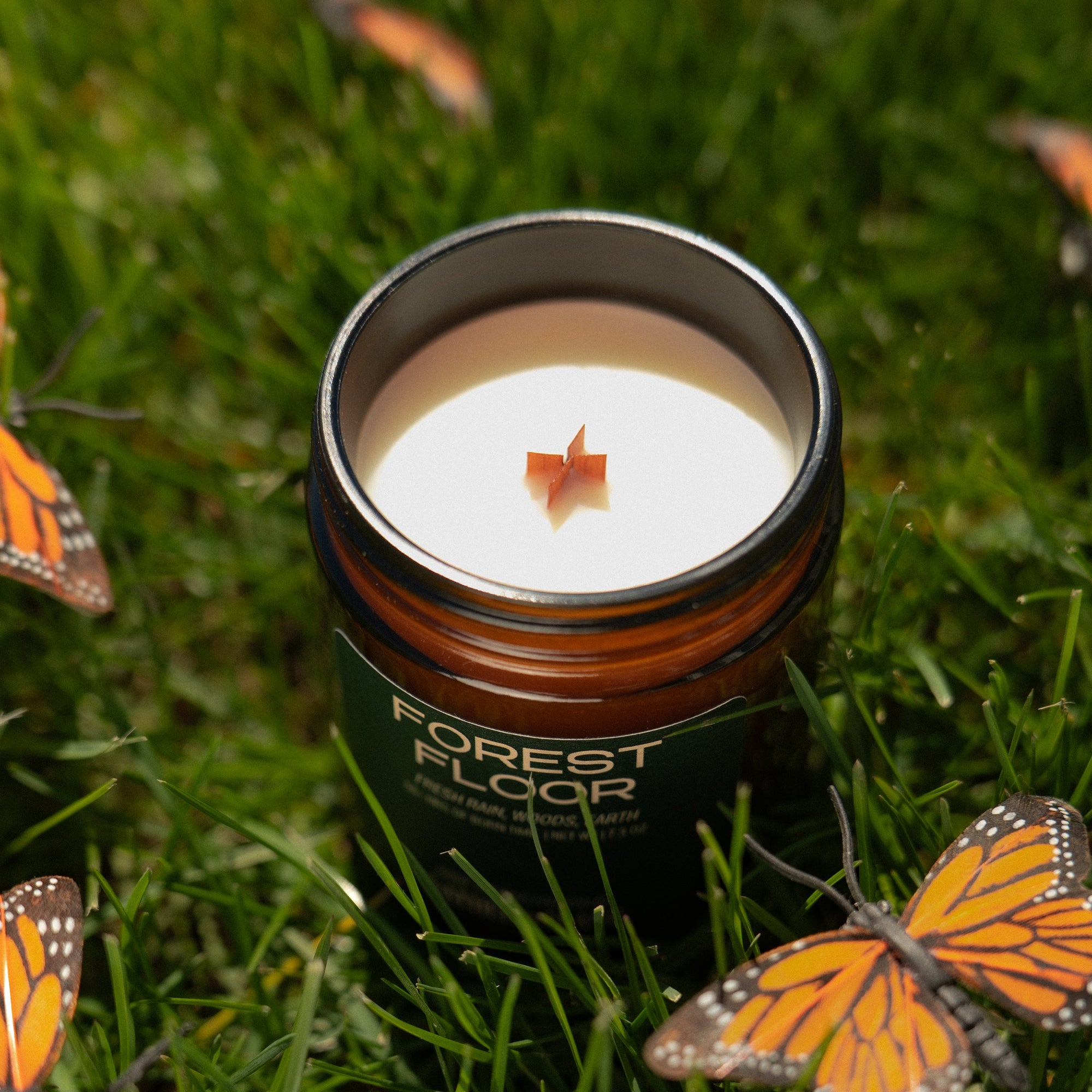 Candle in a container labeled 'Forest Floor' surrounded by butterflies on grass