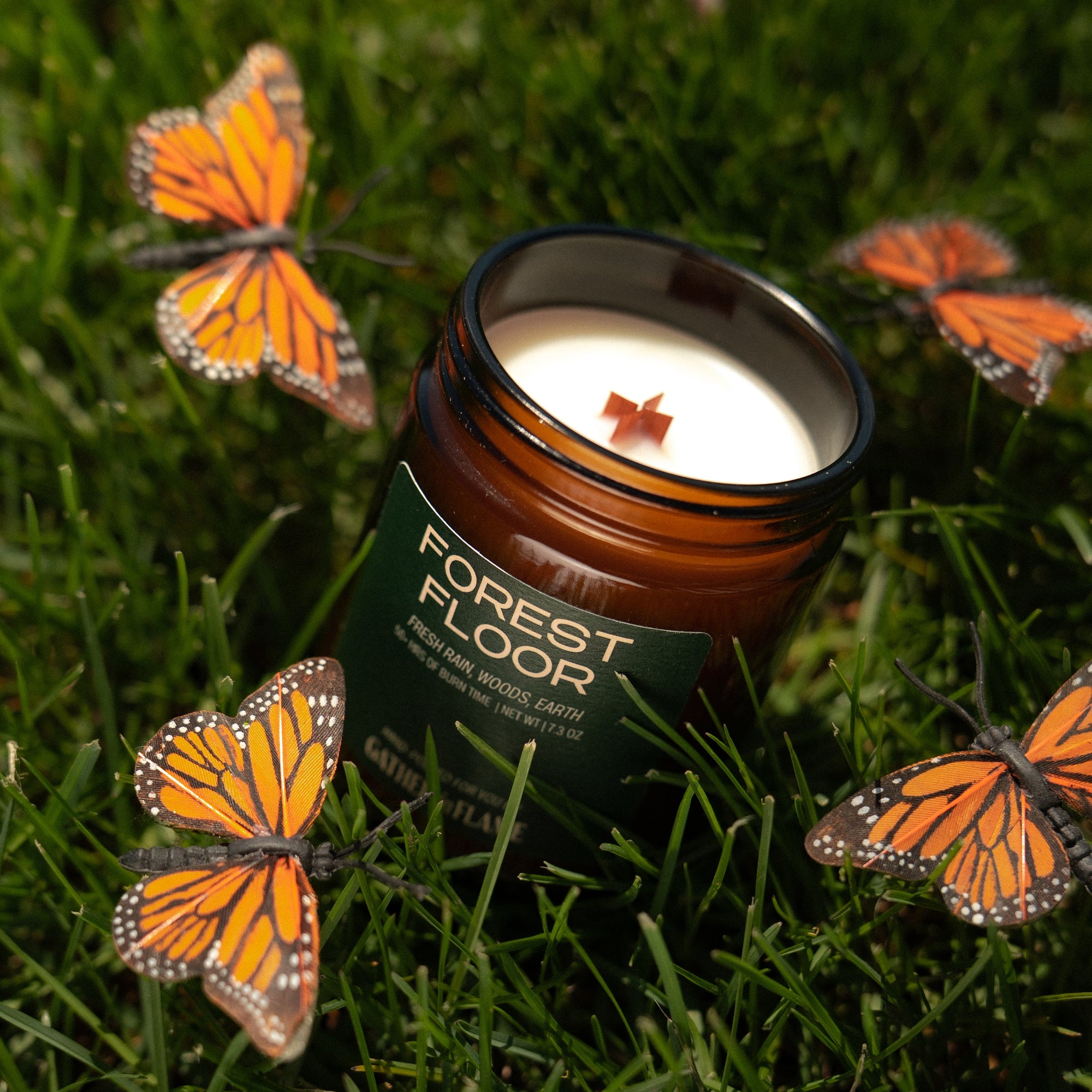 Candle labeled 'Forest Floor' surrounded by butterfly decorations on grass