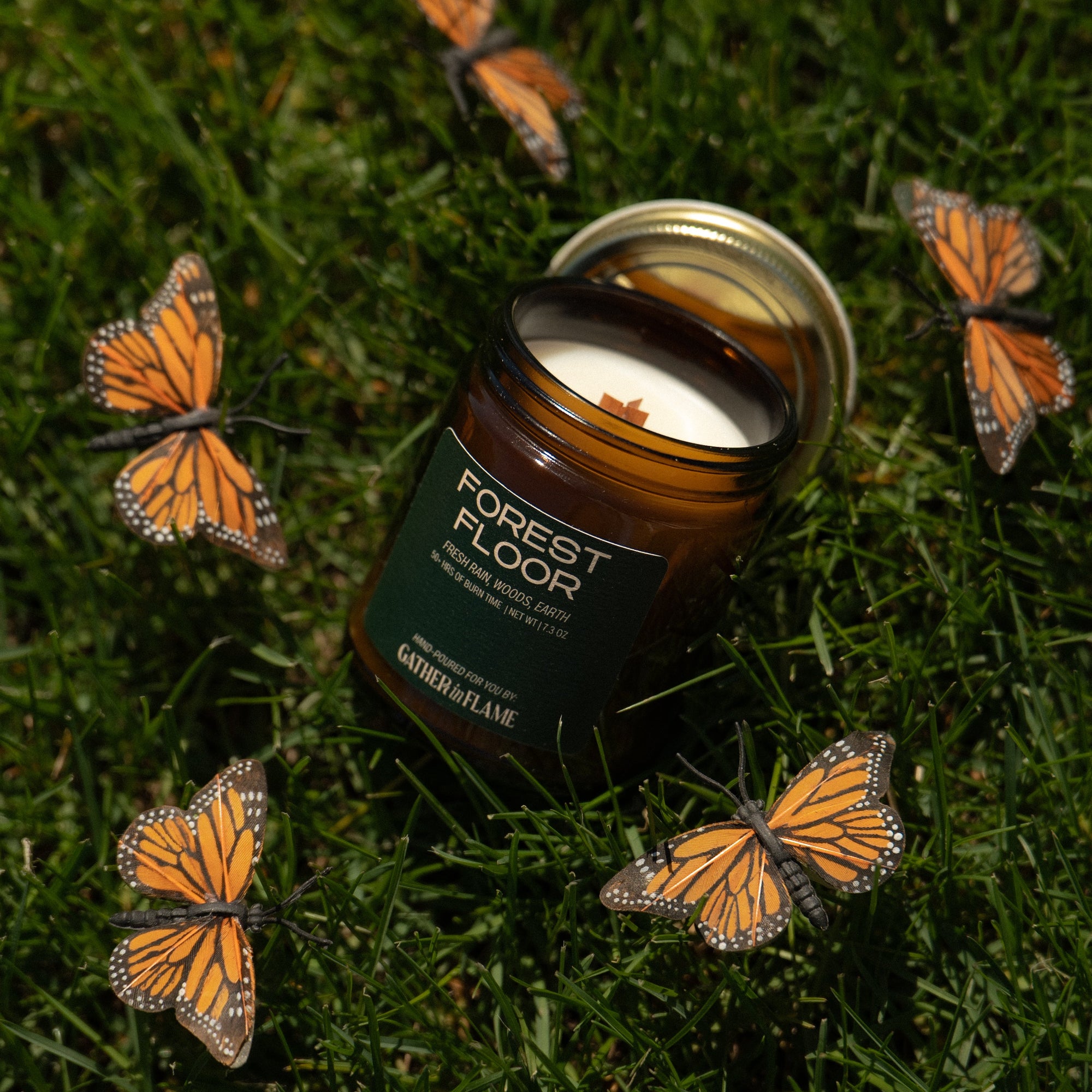 Candle labeled 'Forest Floor' surrounded by monarch butterflies on grass