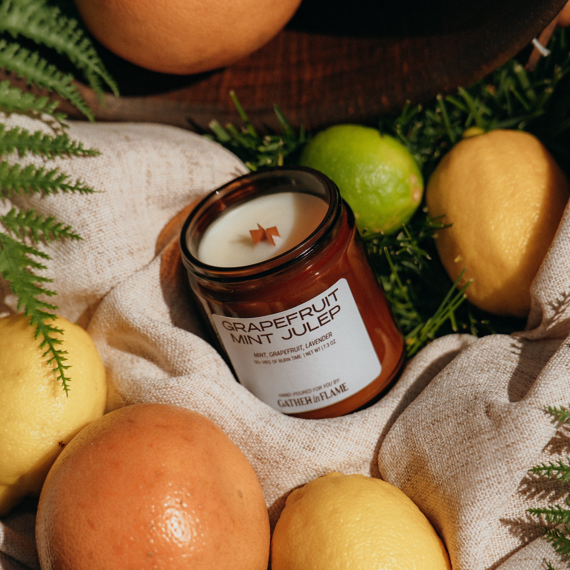 Grapefruit Mint Julep candle surrounded by citrus fruits and greenery on a textured fabric background