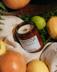 Grapefruit Mint Julep candle surrounded by citrus fruits and greenery on a textured fabric background