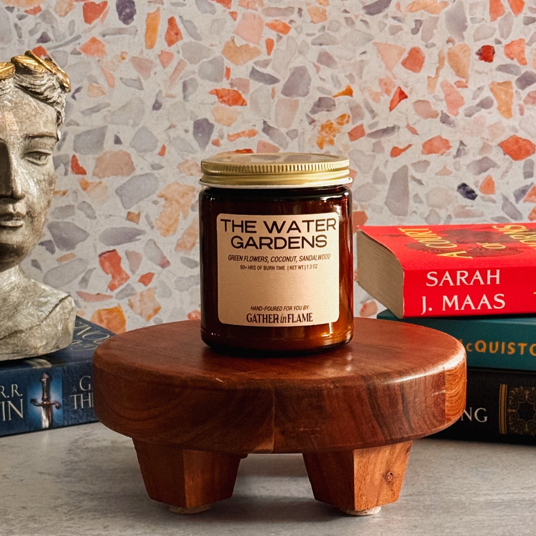 Candle labeled 'The Water Garden' on a wooden stand with books and a bust statue in the background.