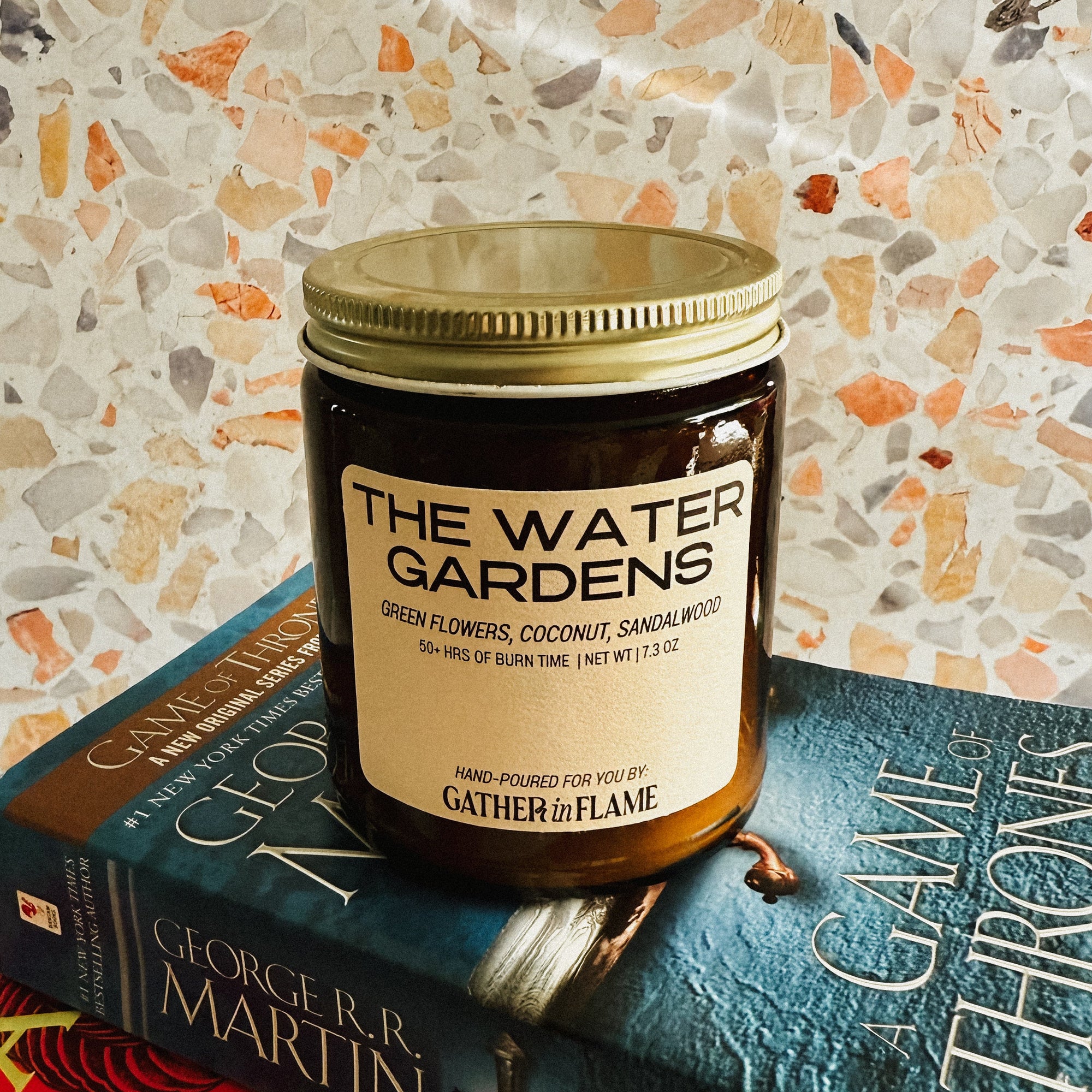 Candle labeled 'The Water Gardens' on top of books with a patterned wall background