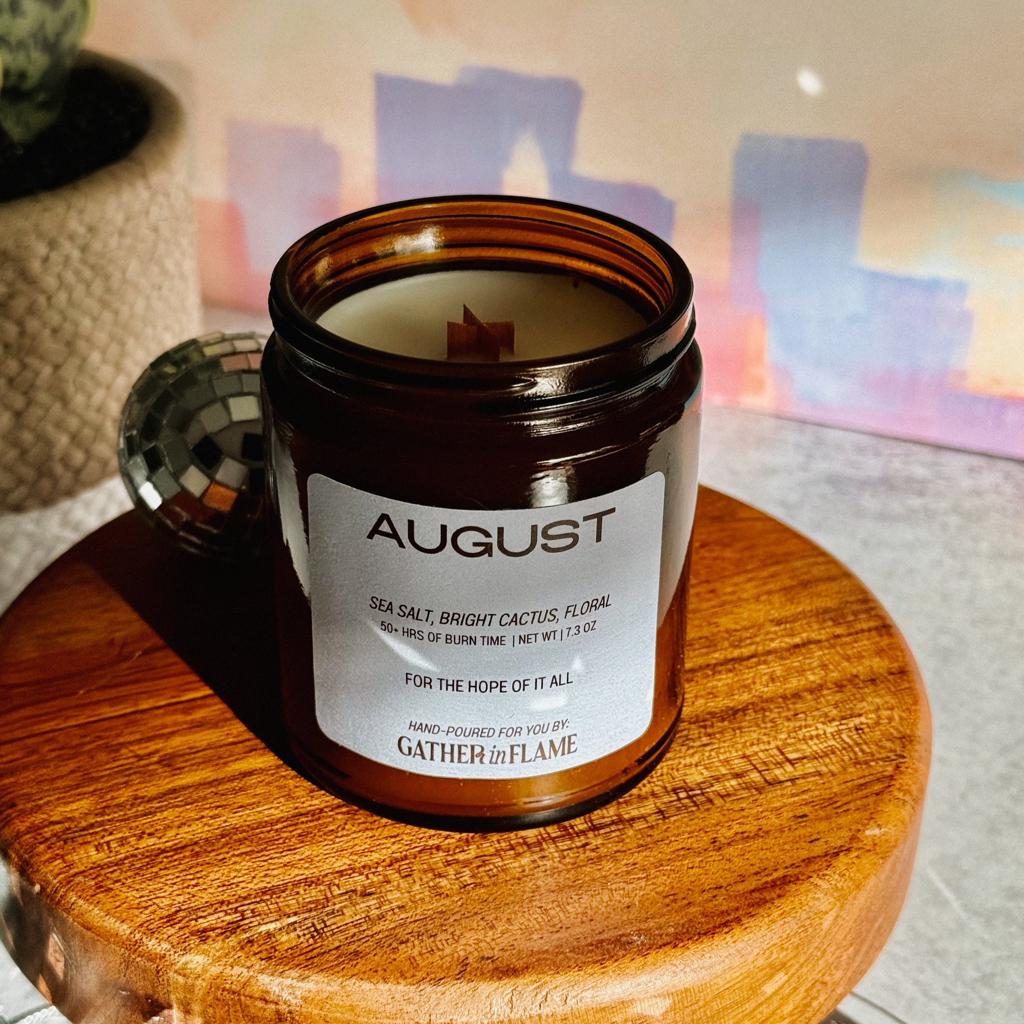 Candle in an amber jar labeled 'August' on a wooden stand with a blurred background.