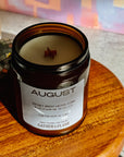 Candle in a jar labeled 'August' on a wooden surface with a colorful abstract painting in the background.