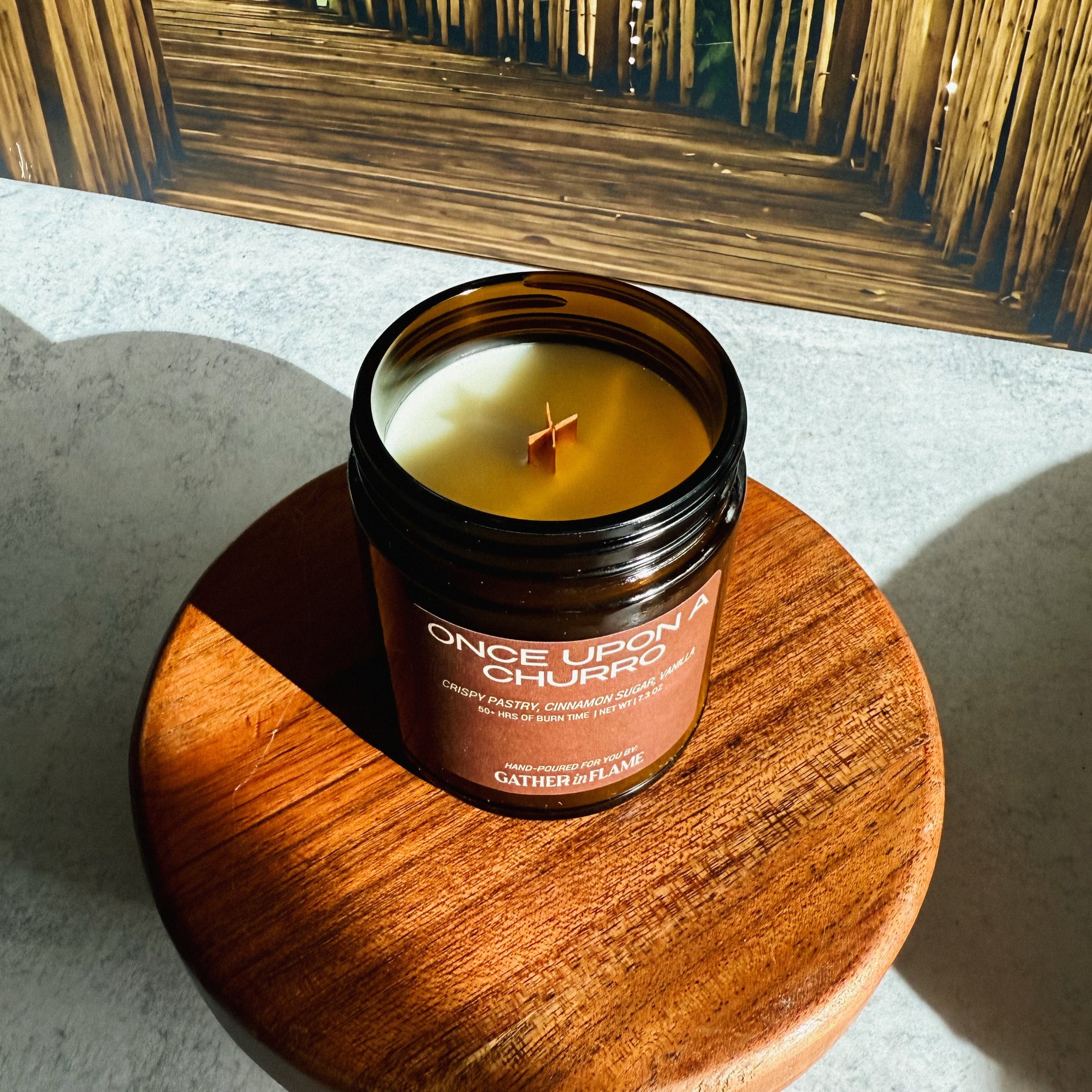 Candle in a glass jar labeled 'Once Upon a Churro' on a wooden coaster with a reflective surface in the background.