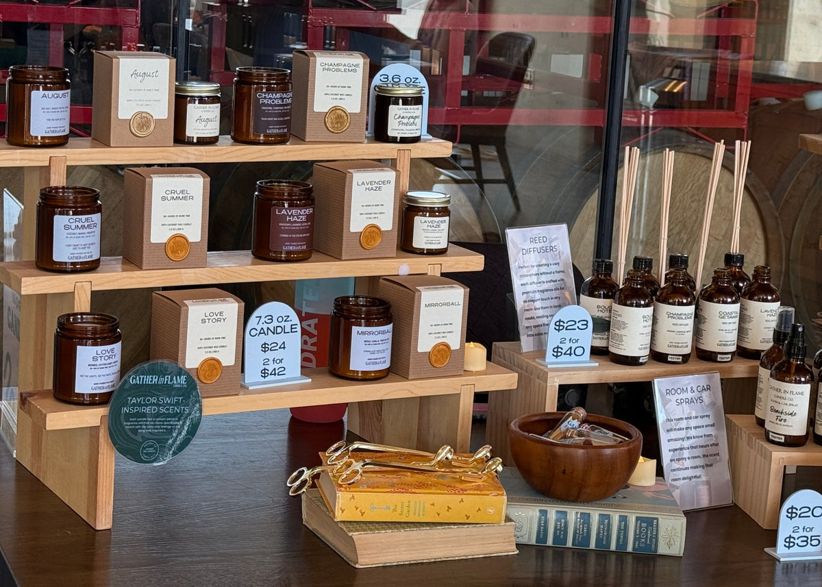 A retail display of artisanal home fragrance products, including candles, diffusers, and essential oils, arranged on wooden shelves and tables inside a shop with large windows and visible wooden barrels in the background.