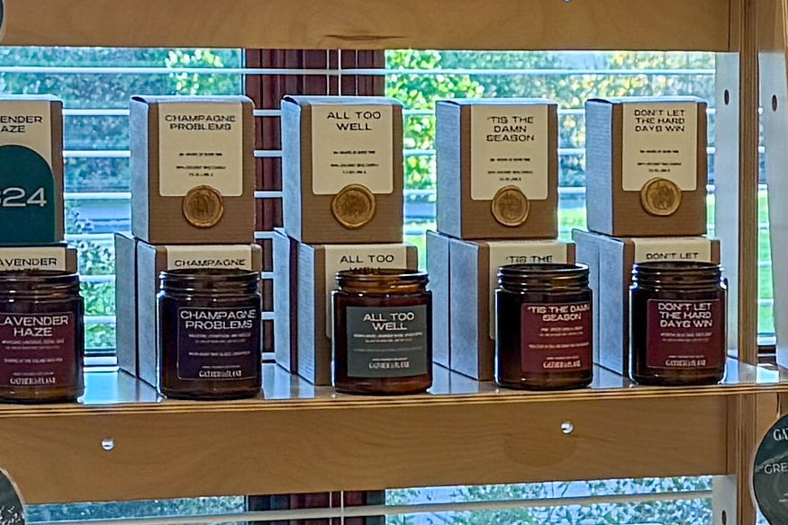 Wooden retail shelves displaying multiple amber glass candle jars with kraft paper boxes and small round boxes, set in front of a window with blinds.