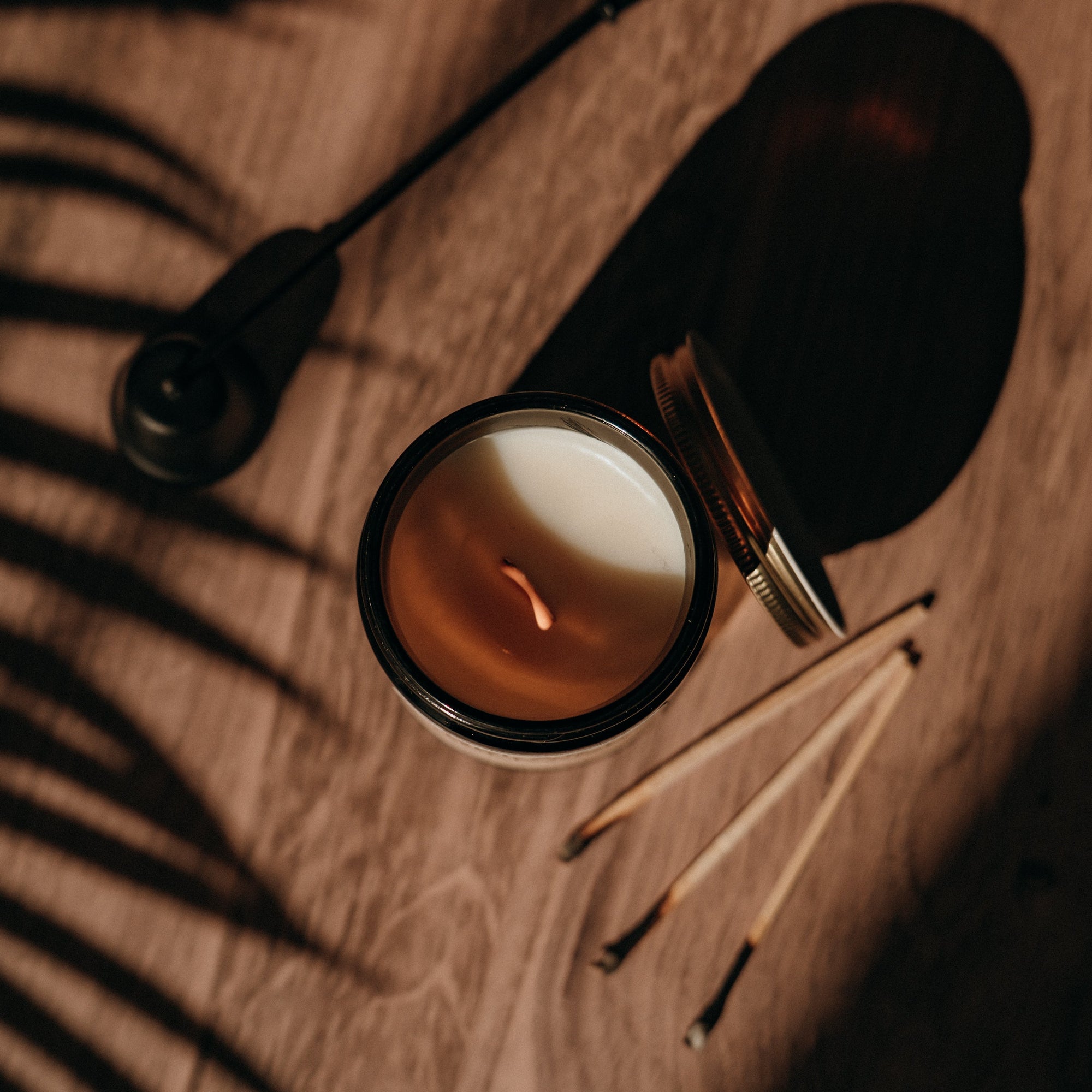 Candle in a glass holder on a wooden surface with a pen and leaf shadows.